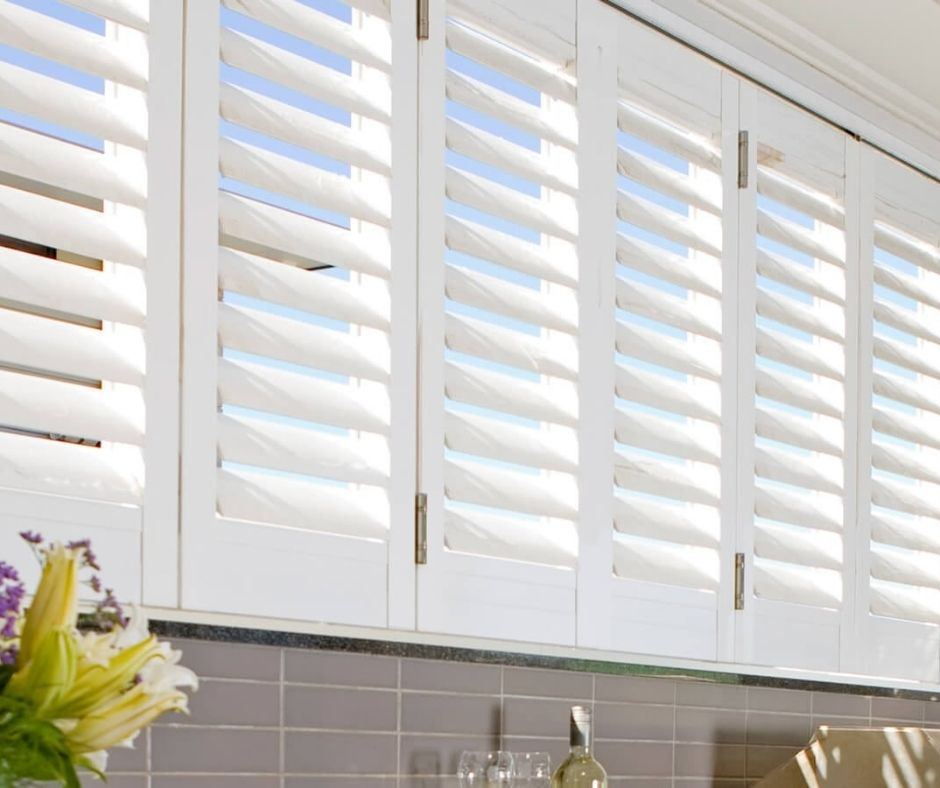 White plantation shutters installed over a kitchen sink with grey subway tile backsplash and natural light.