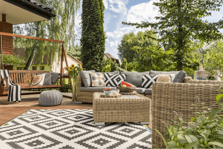 Outdoor patio with woven wicker sofa and chairs, patterned black-and-white rug, wooden deck, and garden greenery, styled with cushions, a coffee table, and a wooden swing seat