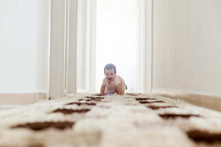 A baby makes its way across the hallway carpet in a brightly lit home.