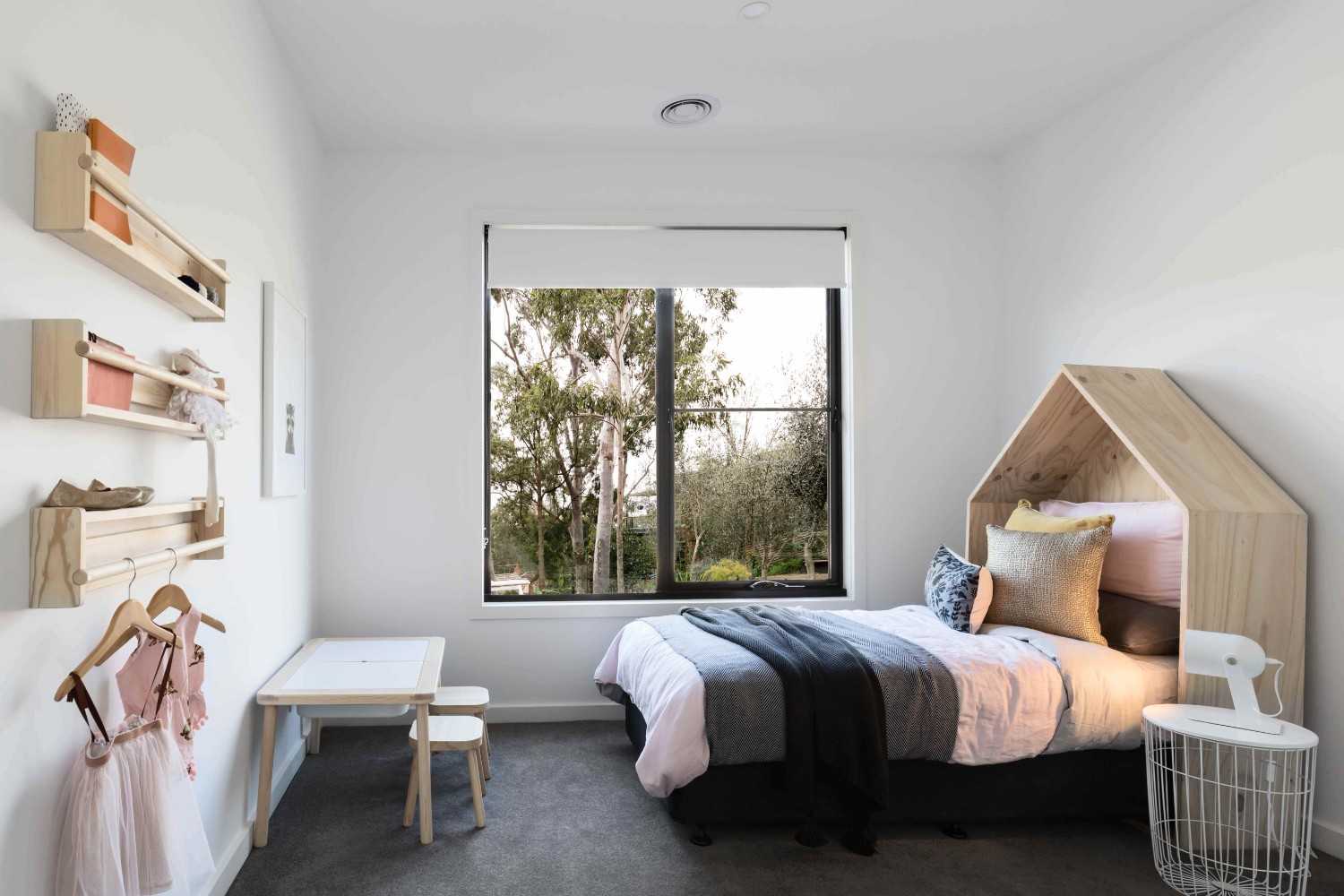 A modern, minimalist children’s bedroom with a single bed featuring a wooden house-shaped headboard, layered pillows, and a white side table with a lamp. A large window fitted with roller blinds lets in natural light and shows trees outside.