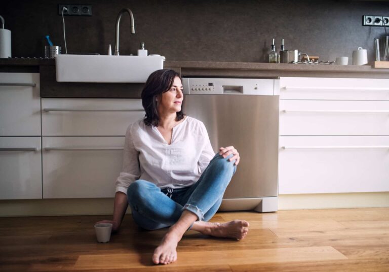 A woman sitting on a timber floor by the kitchen sink.