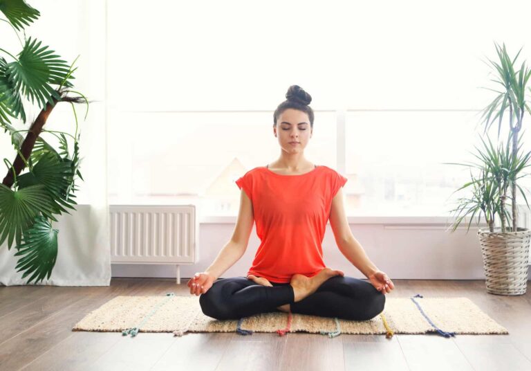 A woman meditating on the floor in a brightly-lit room.