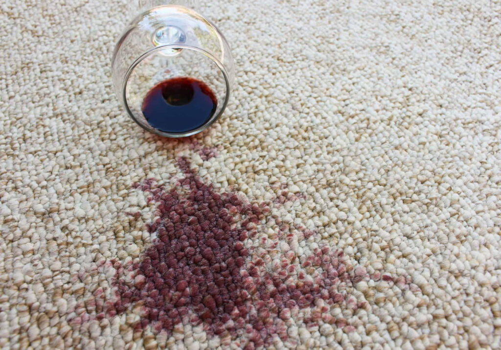 Red wine spilled on a light-coloured wool carpet beside an overturned wine glass.