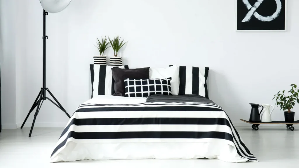 Contemporary bedroom with black and white striped bedding, matching throw pillows, two potted plants with striped planters, tripod floor lamp, small side table with decorative items, and minimal mo...