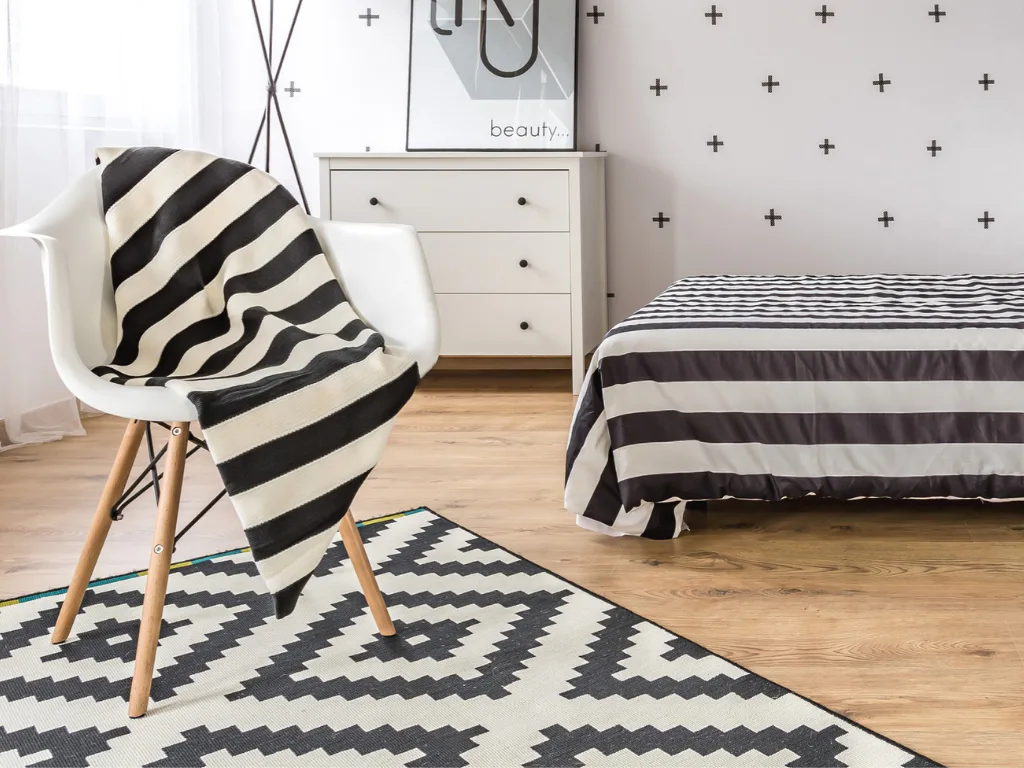 Contemporary bedroom featuring a white mid-century chair with black and cream striped throw blanket, geometric patterned rug, white dresser, and striped bedding against wallpapered accent wall with...