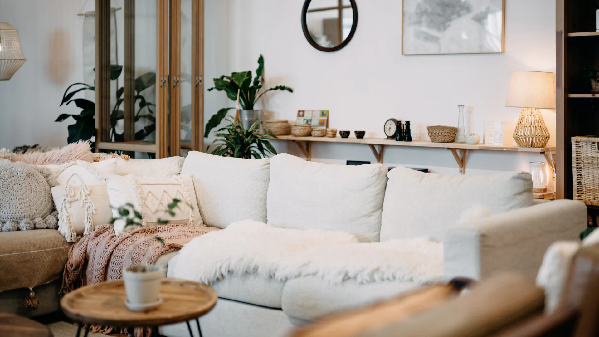 Bright bohemian living room featuring a cream-colored sofa with decorative pillows and knit throw blanket, potted green plants, wooden floating shelves with home decor, round black mirror, and wove...
