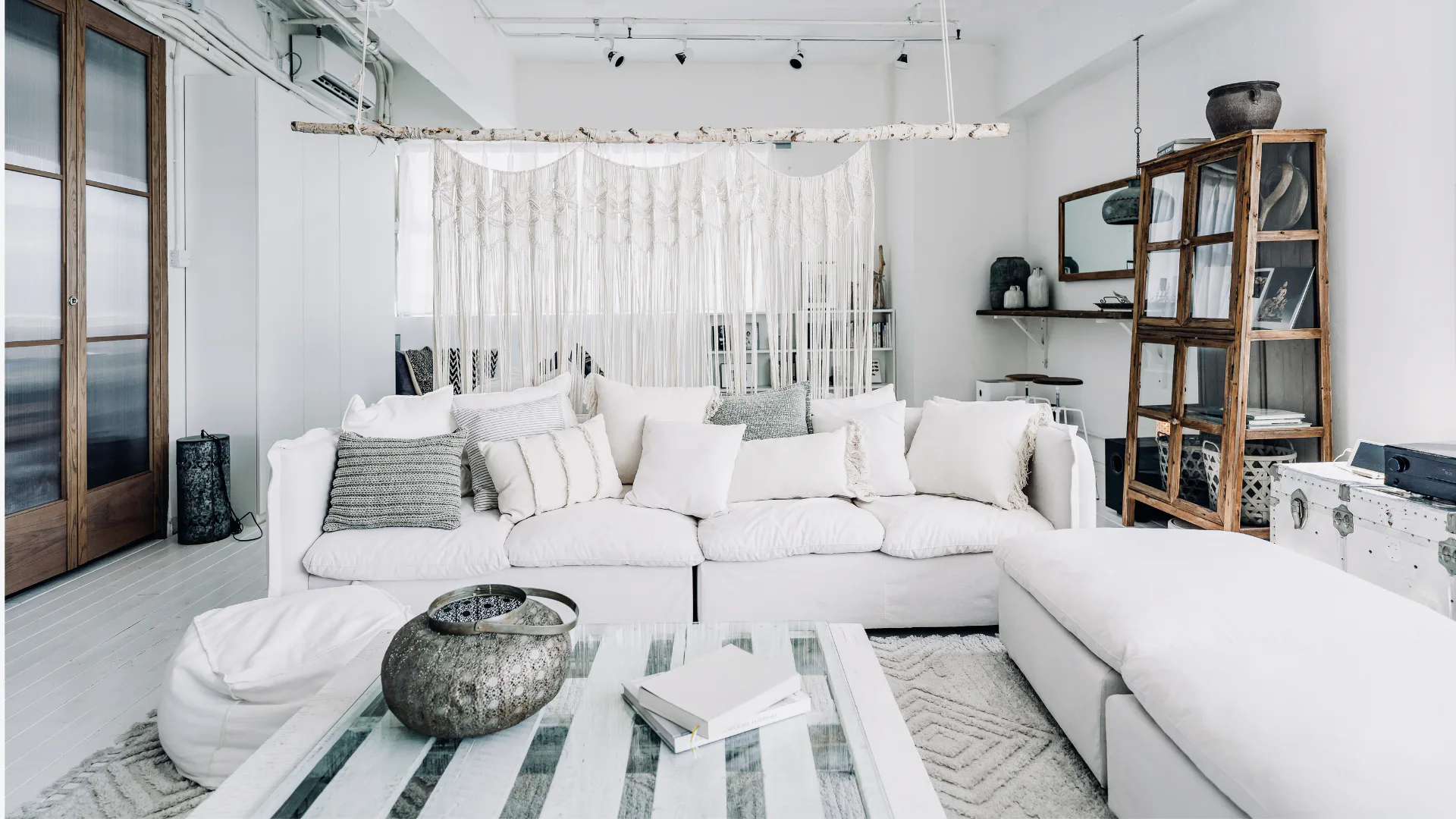 Bright bohemian living room featuring a large white sectional sofa with patterned pillows, macramé wall hanging, wooden glass cabinet, striped area rug, and natural light from tall windows.