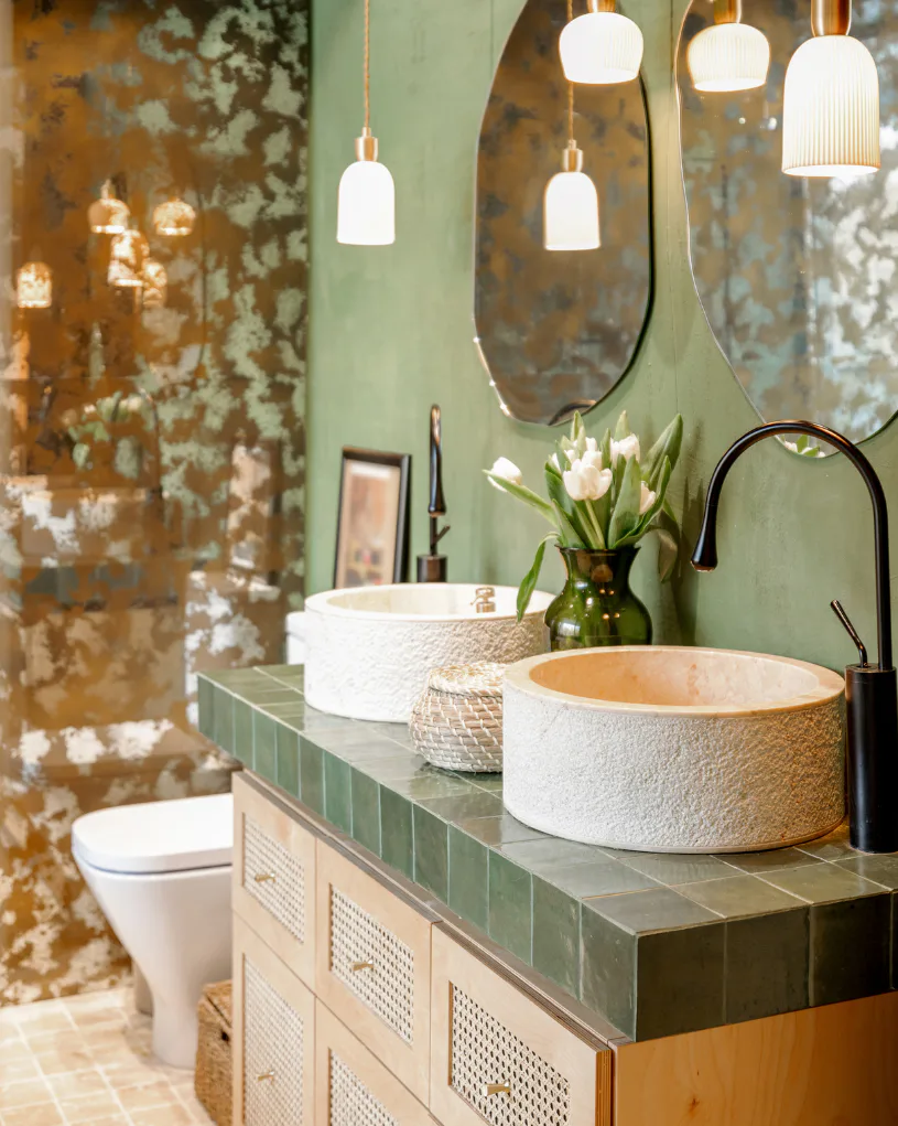 Bohemian bathroom featuring two textured ceramic vessel sinks on a green tile vanity, black faucets, white pendant lights, mirrors, and weathered bronze wall panels with fresh white tulips.