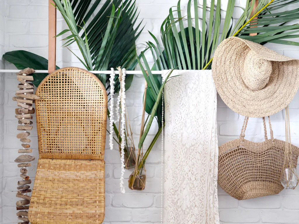 White brick wall decorated with hanging woven baskets, straw hat, dried palm leaves in glass vases, white beaded garland, and woven storage bags creating a botanical home decor display