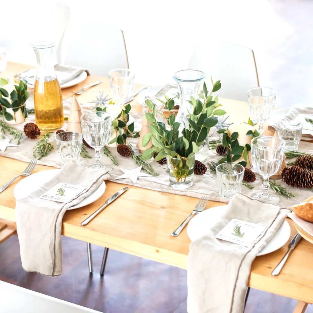 Formal dining table with natural wood surface, white linens, clear glassware, place settings with herb sprigs, eucalyptus and pinecone centerpieces, and golden beverage pitcher.