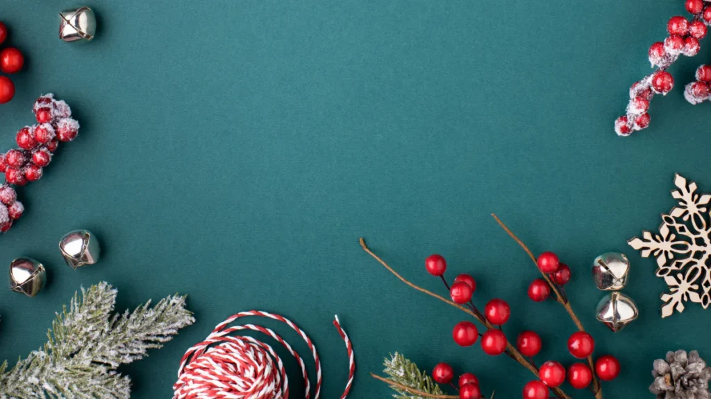 Flat lay holiday composition on teal background featuring frosted red berries, silver jingle bells, wooden snowflakes, candy cane twine, and evergreen sprigs arranged in corners around empty space.