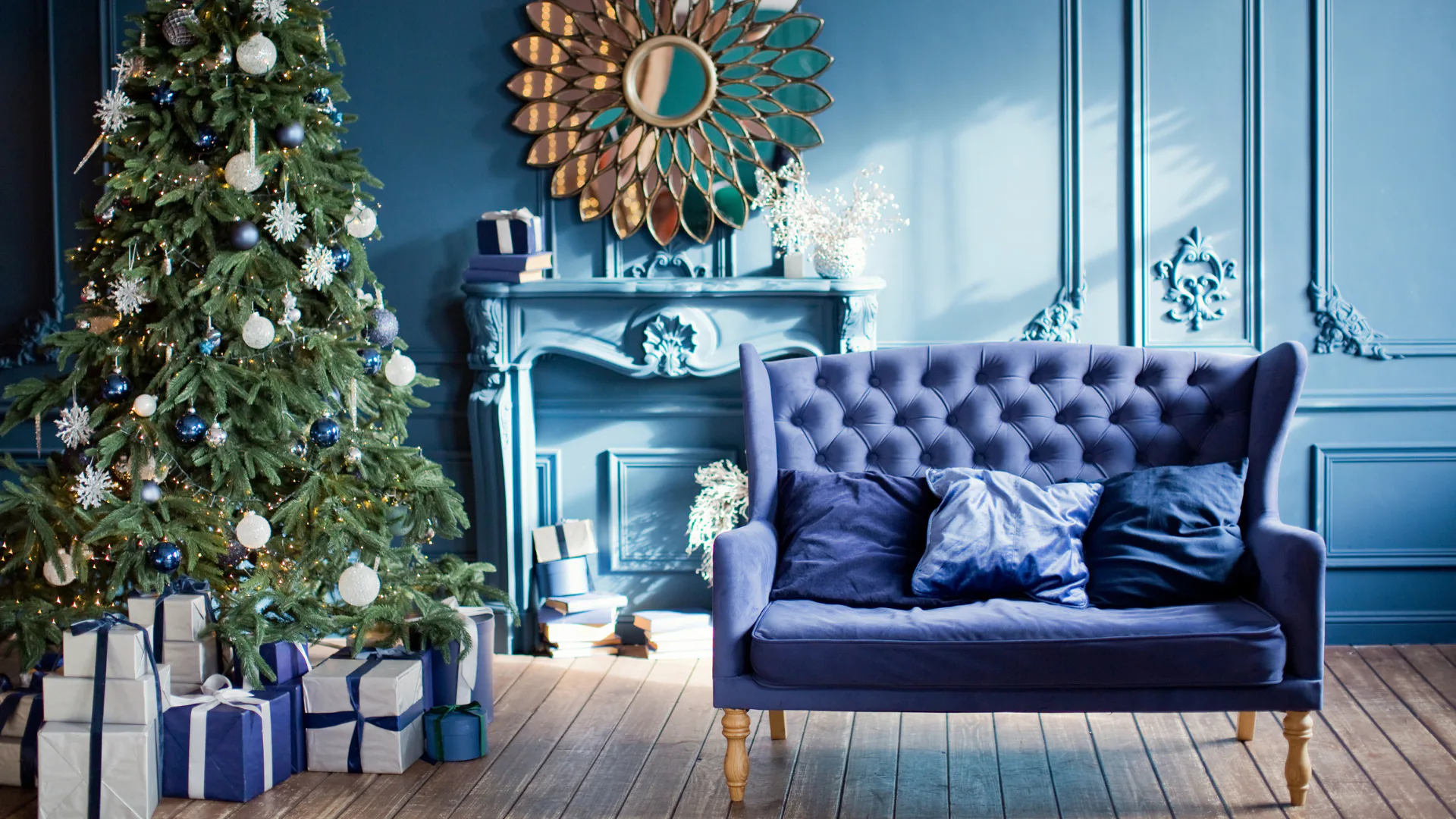 Elegant blue living room featuring a navy tufted sofa, decorated Christmas tree with white and blue ornaments, wrapped presents, ornate fireplace mantel, and decorative sunburst mirror on teal wall.