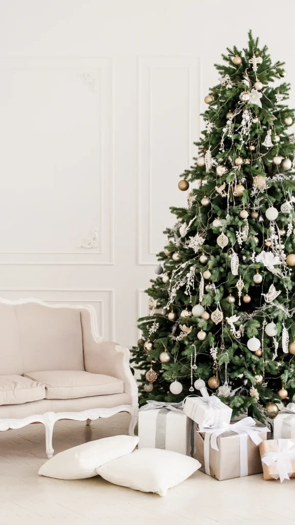 Fully decorated Christmas tree with green branches, gold and white ornaments, white decorative elements, and hanging garland next to cream armchair with wrapped gifts below