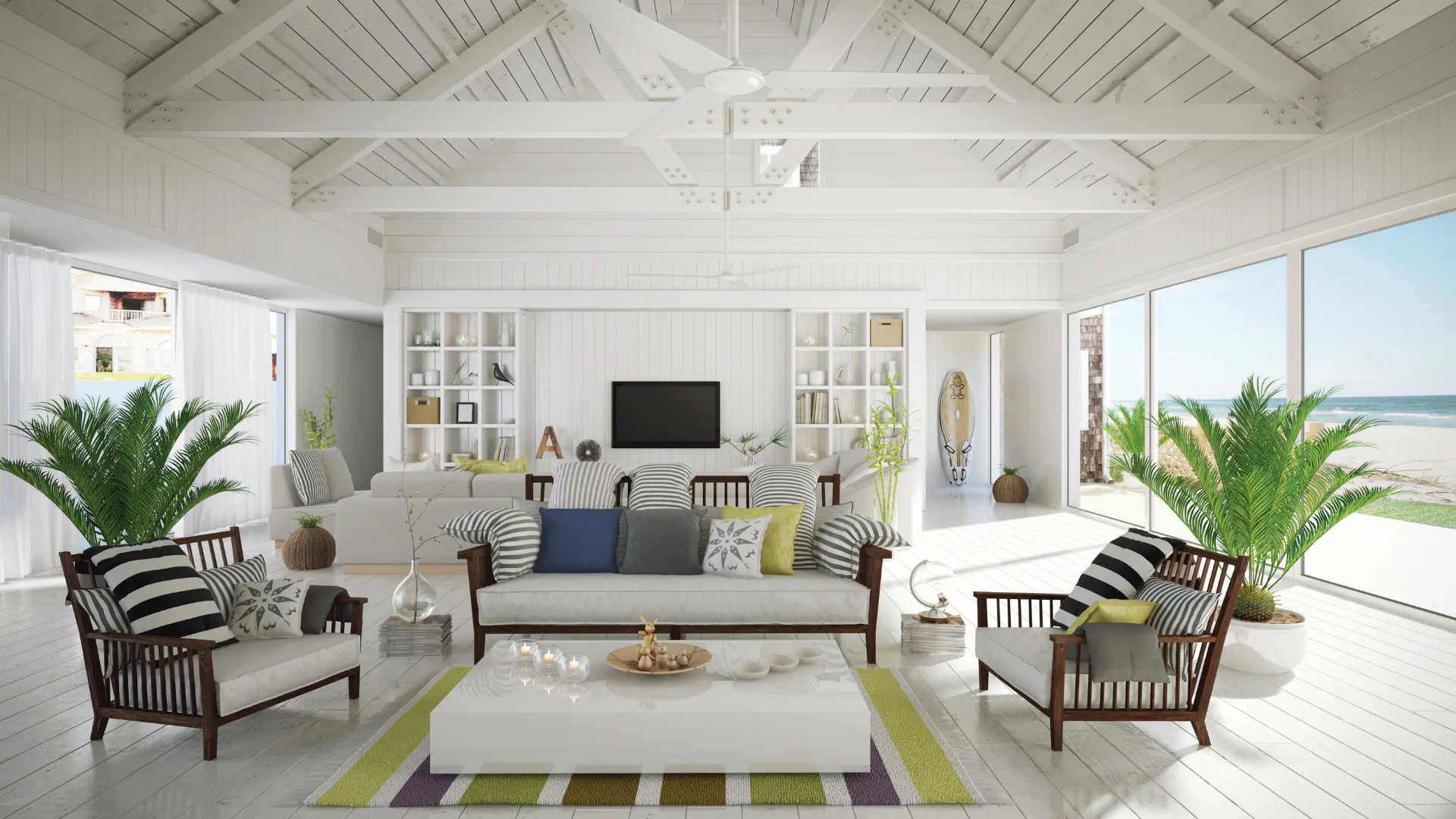 Bright, airy coastal living room with vaulted white ceiling, wooden beams, striped accent chairs, white sofa, palm plants, built-in shelving, ocean view through glass doors, and green-striped area ...