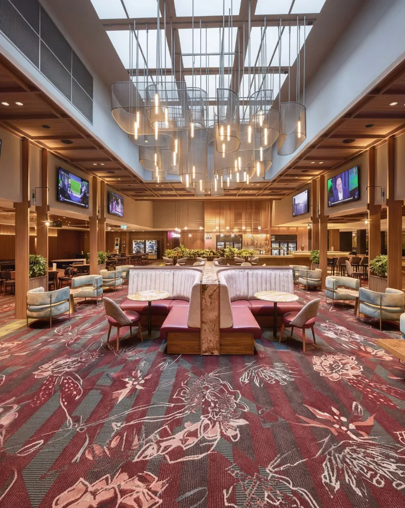 Expansive hotel lobby featuring burgundy and gray patterned commercial carpet with floral design, modern pendant lighting, wood paneling, contemporary seating areas, and central architectural feature.