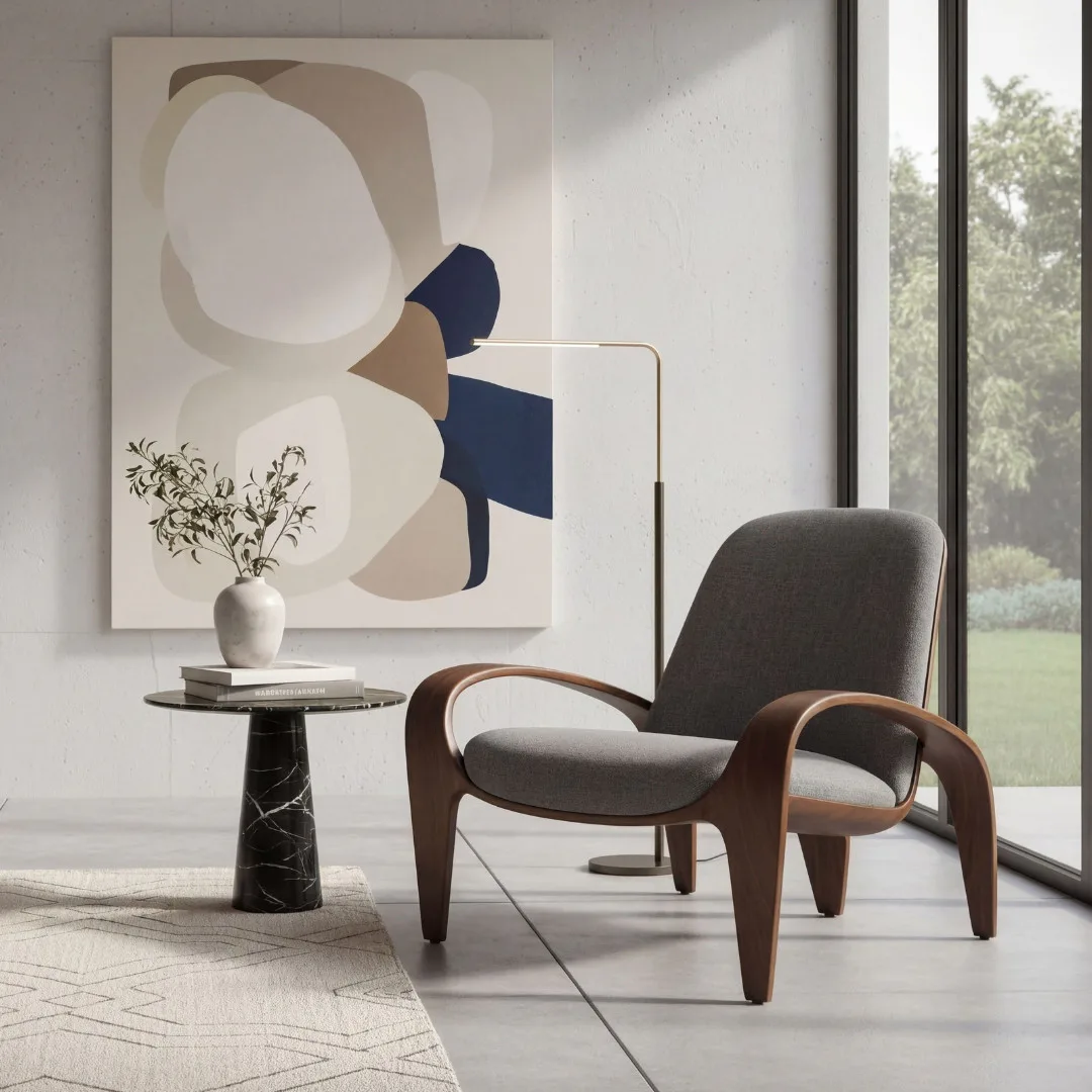 Mid-century modern gray upholstered lounge chair with curved wooden arms beside a black marble side table holding a white vase with dried flowers, beneath abstract geometric wall art