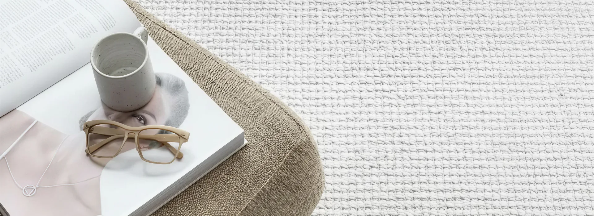 Overhead view of a neutral-toned living space featuring a beige textured area rug, white throw blanket, ceramic mug, eyeglasses, open magazine, and pastel accessories on white surface.