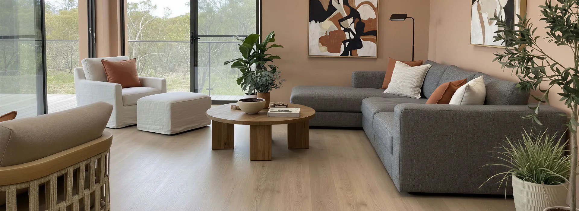 Contemporary living room featuring light hybrid flooring, gray sectional sofa with orange and cream pillows, white armchair, wooden round coffee table, large windows overlooking trees, and potted p...