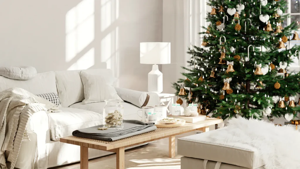 Bright living room with cream sofa, wooden coffee table, white table lamp, and decorated Christmas tree featuring gold bells, white hearts, and gold ornaments by sunlit windows.