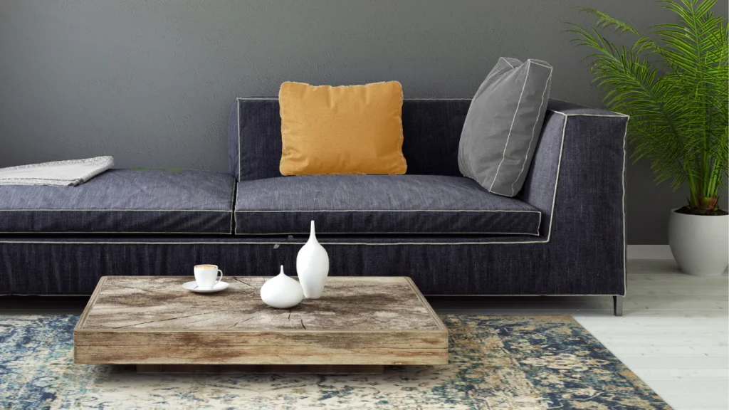 Dark gray upholstered sectional sofa with white piping, mustard yellow and gray pillows, wooden coffee table with white ceramic vases, patterned area rug, potted palm plant, and gray textured wall.