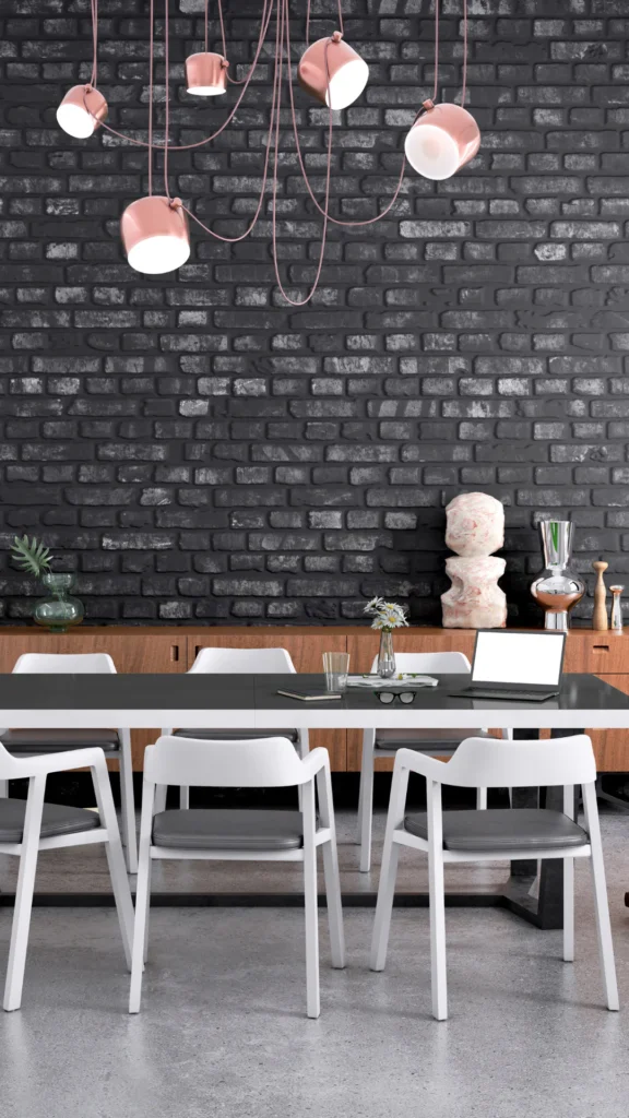 Modern office workspace featuring five rose gold pendant lights with white interiors hanging from coral cables against a dark brick wall, with white chairs and wooden desk below.
