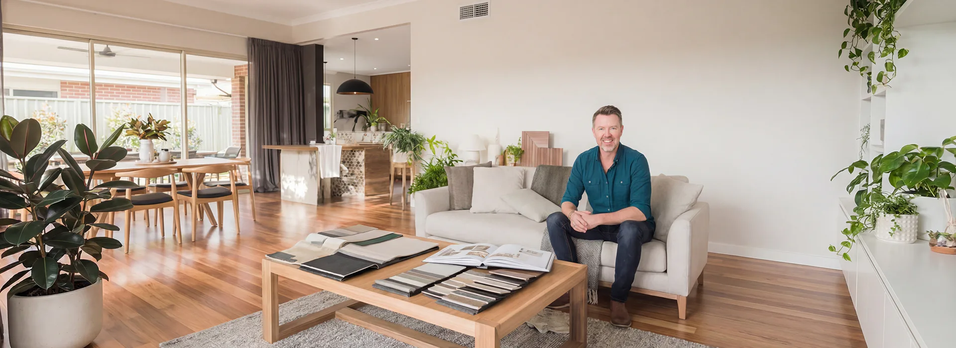 Smiling interior designer in teal shirt sitting on beige couch in modern living room with wooden floors, plants, dining area, and material samples on coffee table