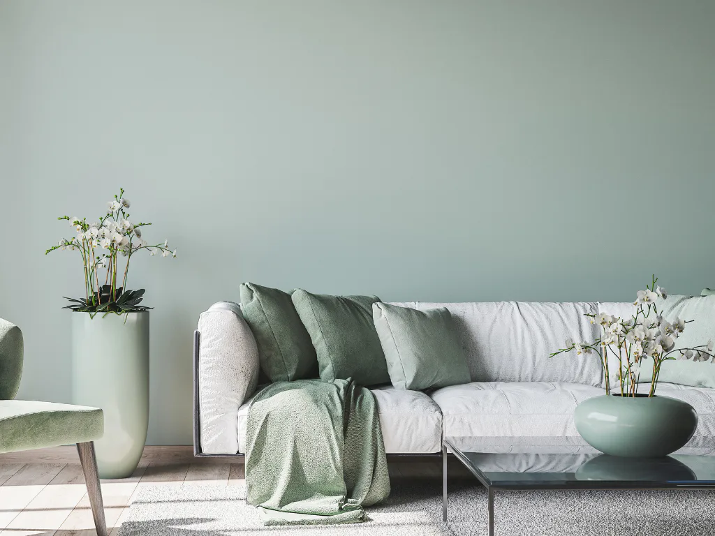 Contemporary living room featuring a light gray upholstered sofa with green and white throw pillows, a green blanket draped over the arm, white potted flowers on side tables, and minimalist decor a...