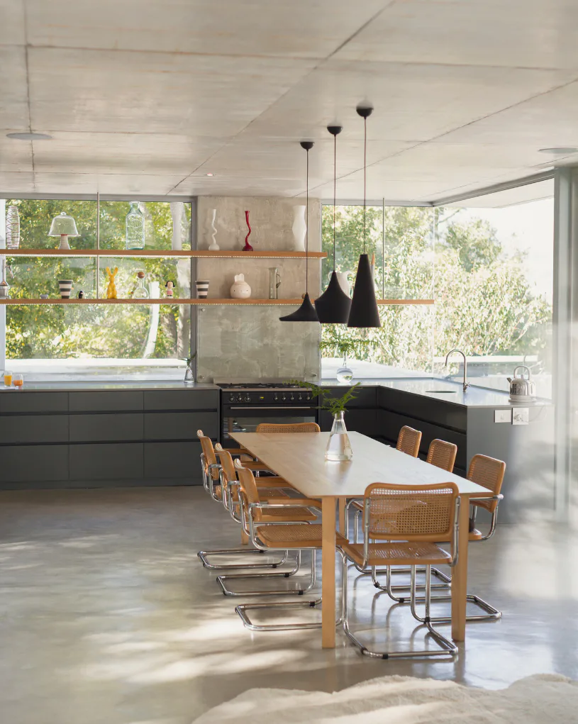Contemporary kitchen-dining space with black pendant lights above wooden table, natural wood chairs with caning, dark cabinetry, open shelving, large windows overlooking greenery, and polished conc...