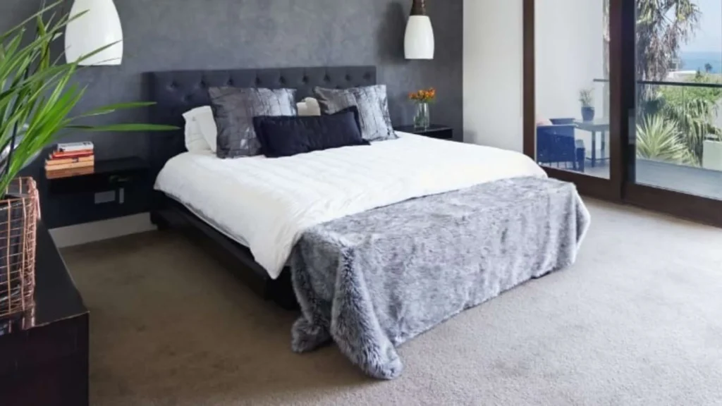 Contemporary bedroom with dark upholstered headboard, white and gray bedding, pendant lights, potted plants, and floor-to-ceiling windows overlooking ocean and palm trees