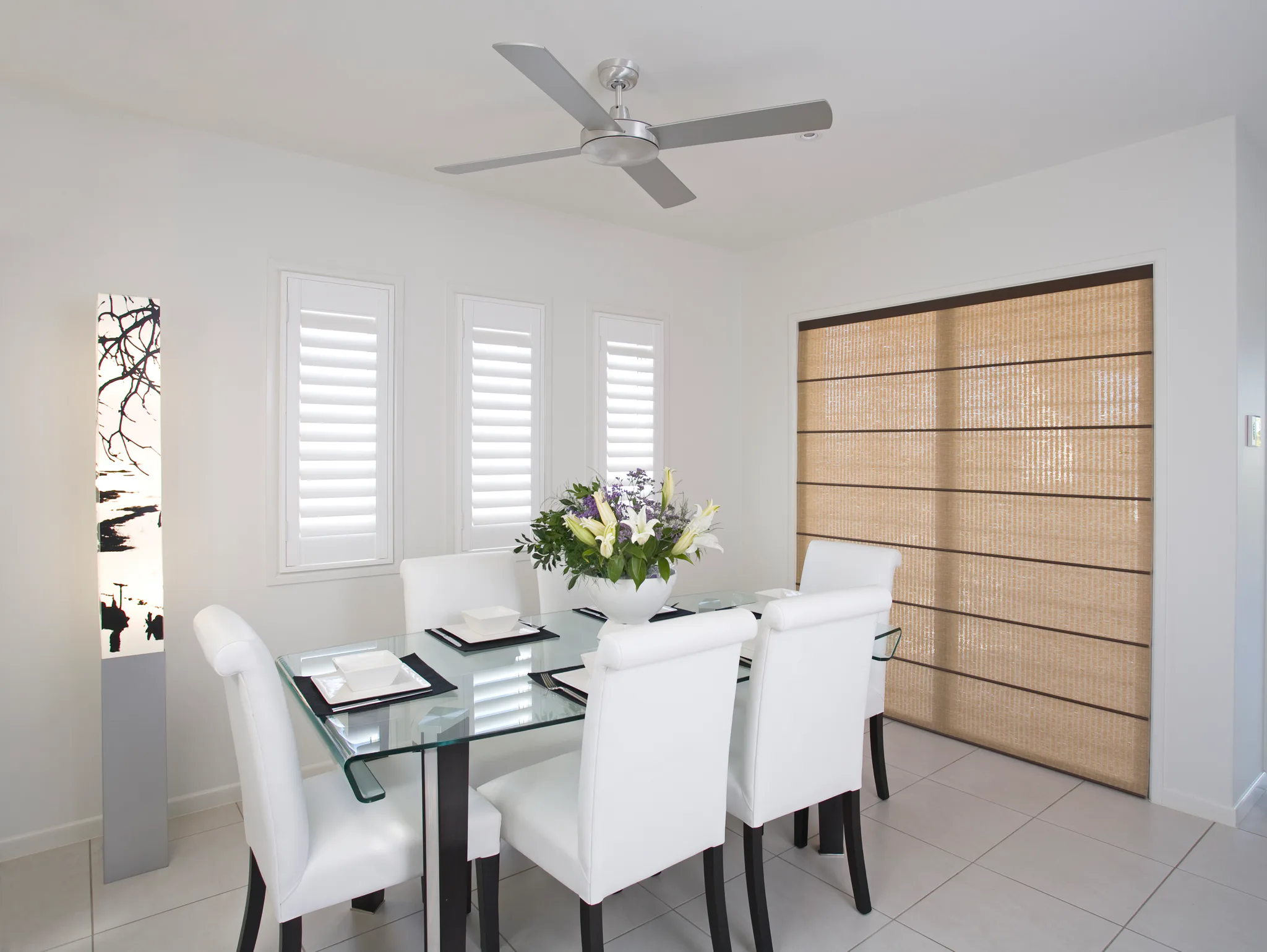 Contemporary dining room featuring light gray tile flooring, glass table with white chairs, white shutters, ceiling fan, and woven Roman shades on large windows