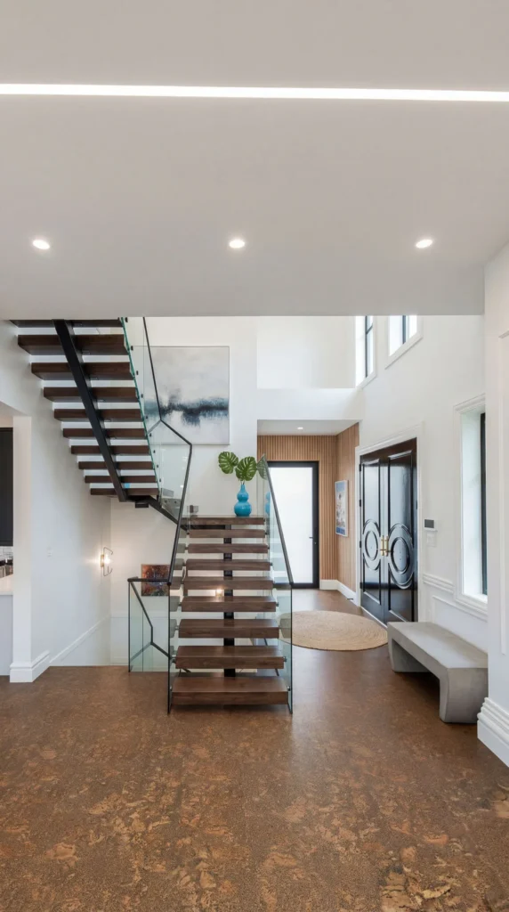 Contemporary foyer featuring a floating wooden staircase with glass railing, cork flooring, white walls, recessed lighting, and a blue vase with greenery on a floating shelf.