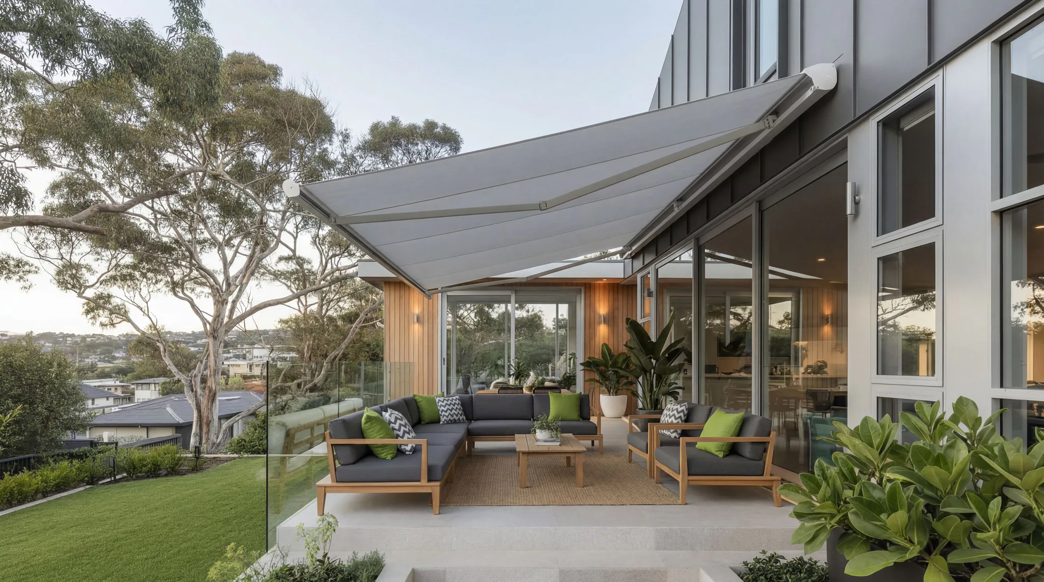 Contemporary outdoor patio featuring a white retractable awning extending over gray upholstered furniture with green accent pillows, surrounded by lush plants and eucalyptus trees