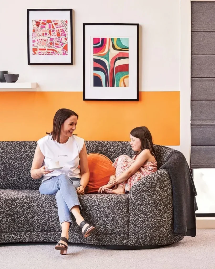 Woman and young girl sitting together on a charcoal textured sofa against a bright orange accent wall, with framed colorful artwork displayed above them