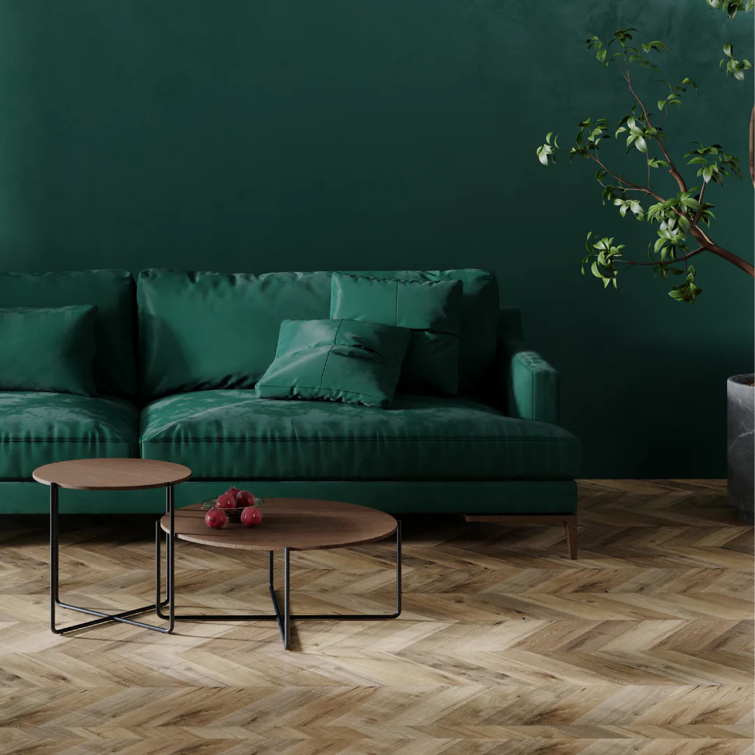 Contemporary living room with deep emerald green sofa, wooden coffee tables with red apples, herringbone wood flooring, and natural branch with green leaves against solid green wall