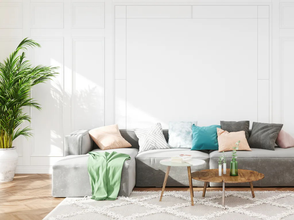 Bright modern living room featuring a gray sectional sofa with colorful throw pillows, wooden coffee table, green throw blanket, potted plant, white shag rug, and paneled white walls with natural s...