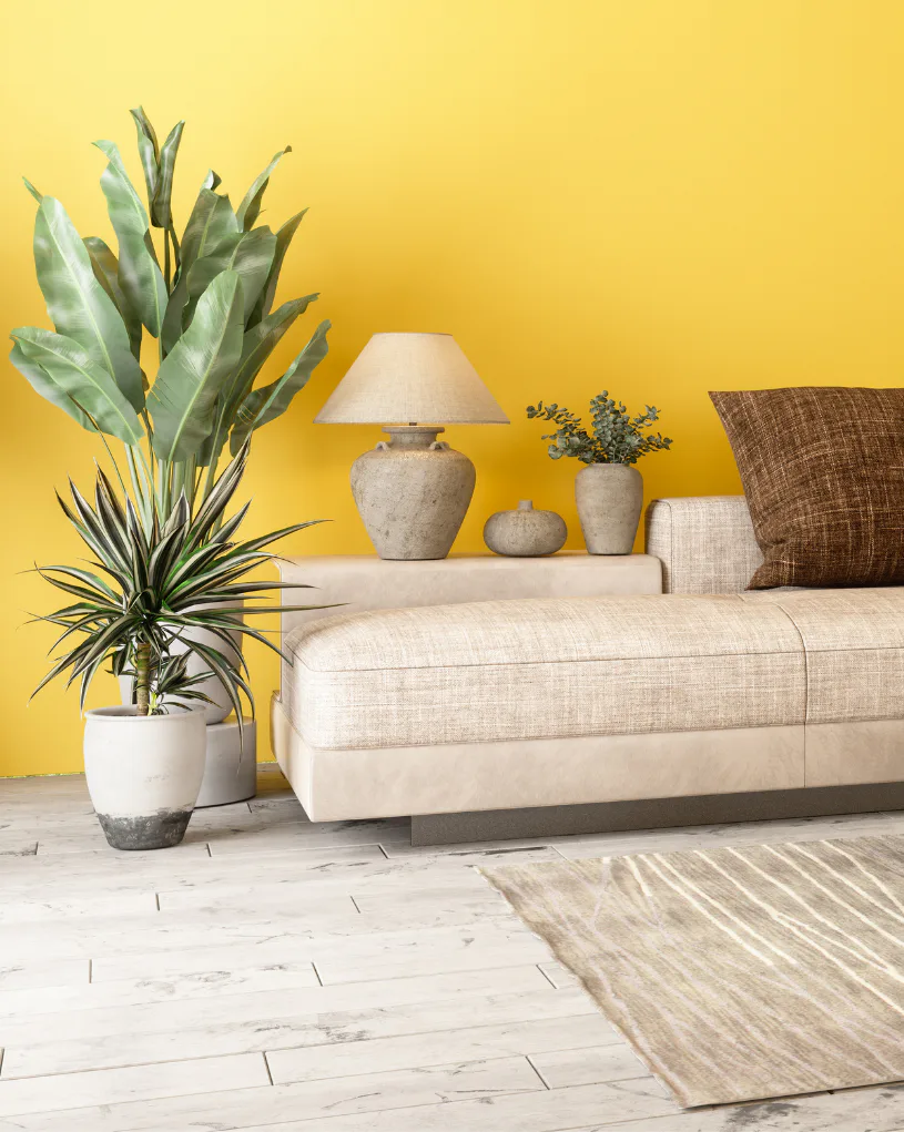 Minimalist living room featuring a cream linen sofa with brown pillows against a bright yellow wall, decorated with potted plants including a tall dracaena in a white planter, ceramic vases with gr...