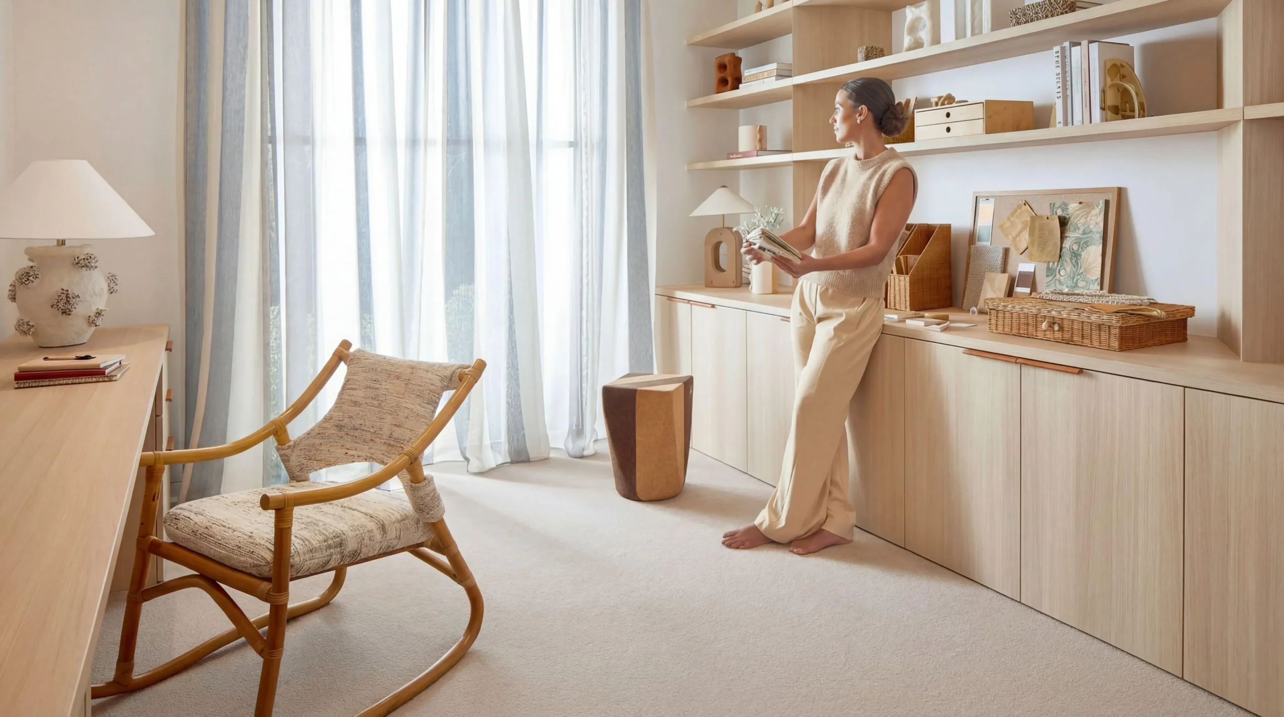 Woman in a beige outfit standing in a modern home office with light wood shelving and serene natural light.