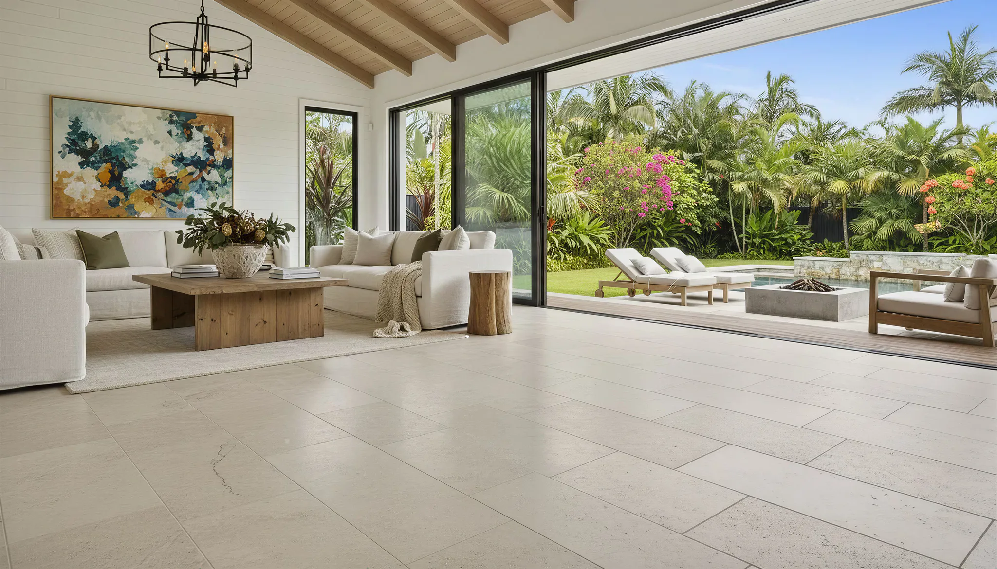 Spacious open-plan living room with cream upholstered furniture, wooden coffee table, and large sliding glass doors opening to a tropical garden patio with loungers and fire pit.