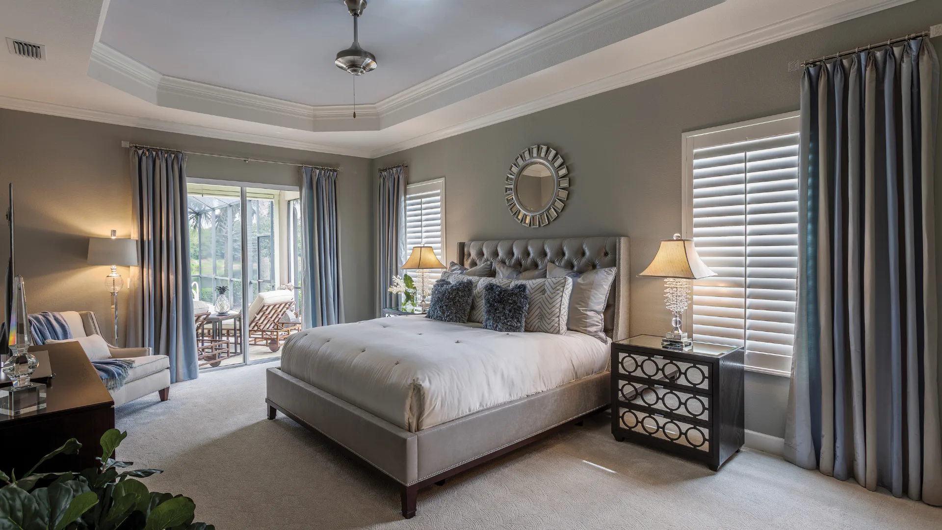 Elegant traditional bedroom featuring a tufted gray upholstered bed, white bedding with decorative pillows, dual nightstands with brass lamps, ornate mirror above headboard, blue drapes, shutters, ...