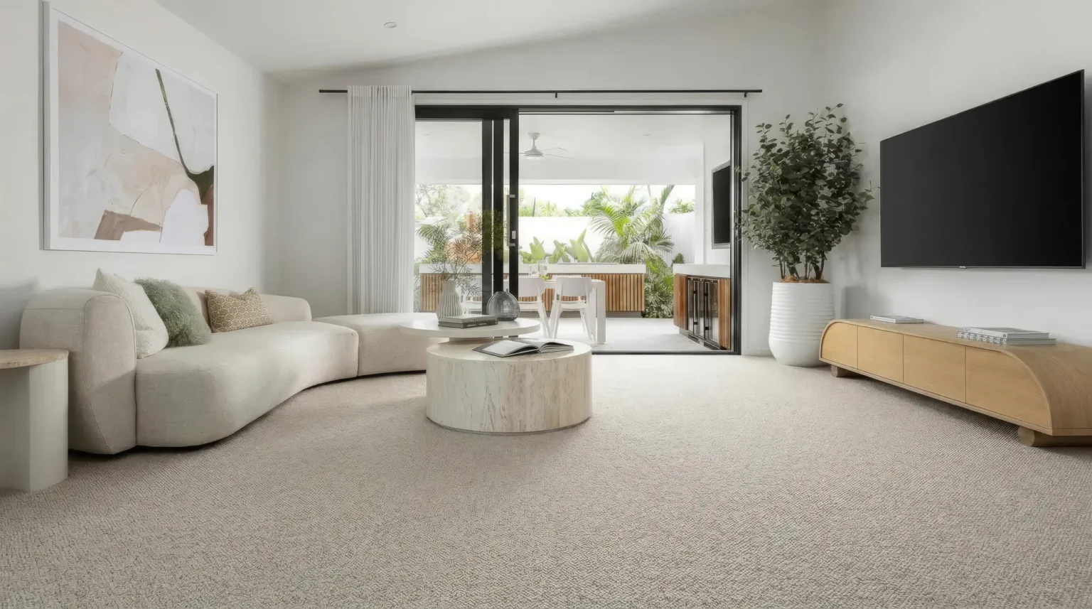 Spacious modern living room featuring a curved beige sofa, round marble coffee table, light gray carpet flooring, wooden media console, potted plants, and floor-to-ceiling glass doors opening to an...
