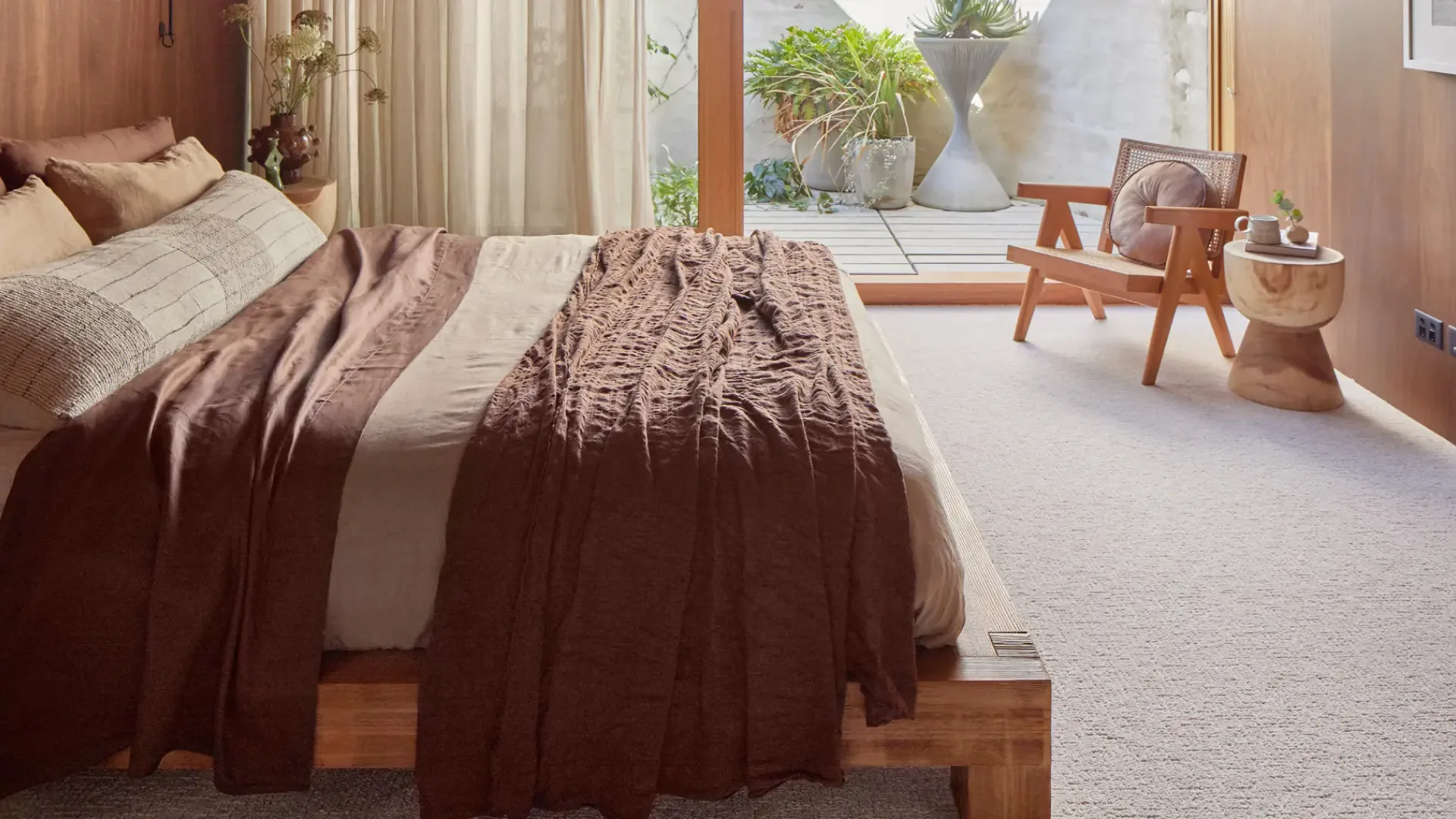 Spacious bedroom featuring a bed with brown and cream layered bedding and textured throw blanket, overlooking an outdoor patio with potted plants and modern furniture on triexta carpet flooring.