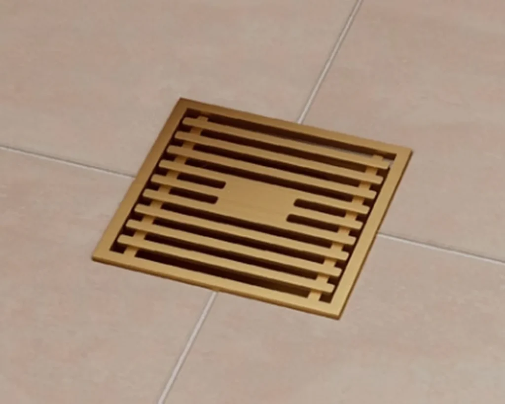Square brass-colored bathroom waste grate with horizontal slats and central rectangular opening, mounted on white drop ceiling tiles with visible grout lines.