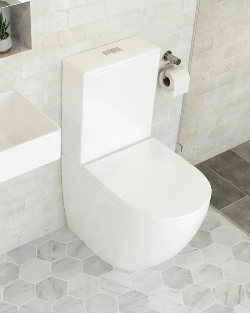 White ceramic toilet with closed seat and lid, mounted against light grey subway tile walls in a contemporary bathroom with marble hexagonal floor tiles and chrome accessories.