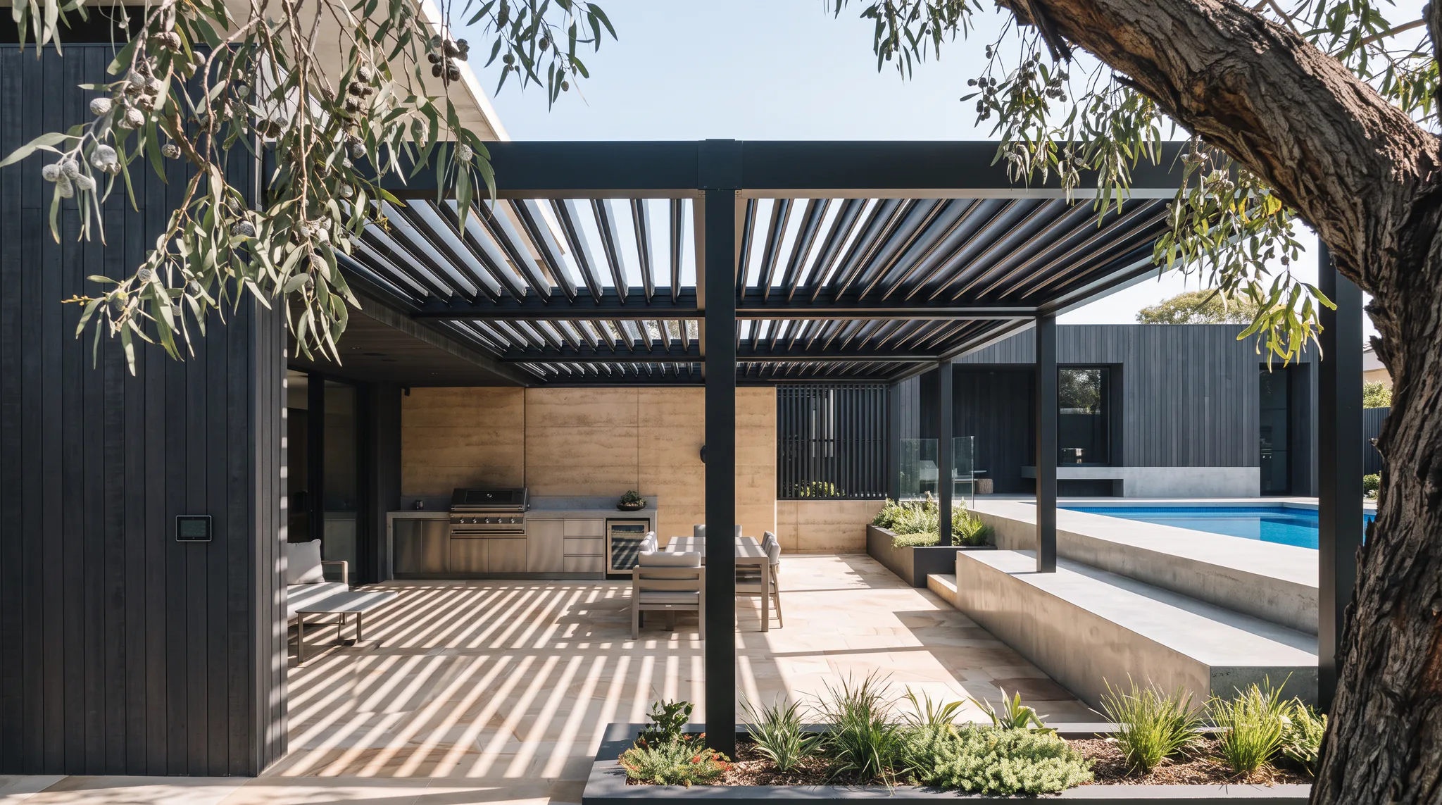 Contemporary black metal pergola with adjustable louver roof covering an outdoor kitchen with stainless steel appliances, dining area, and view of swimming pool beyond
